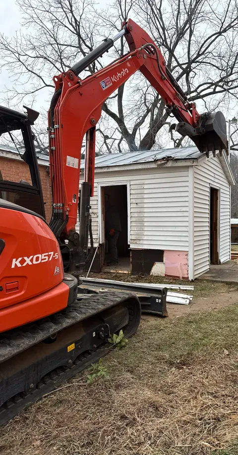 Kubota excavator pulling apart a small white outbuilding.