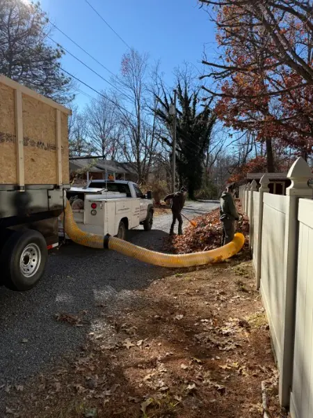 Two crew members vacuuming leaves along a white fence into a dump trailer.